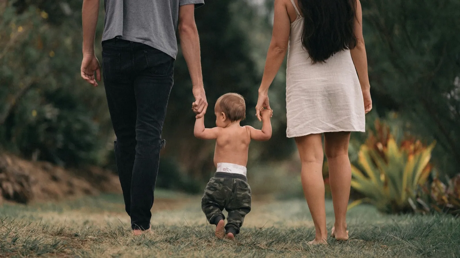 Two parents holding the hands of their baby learning to walk