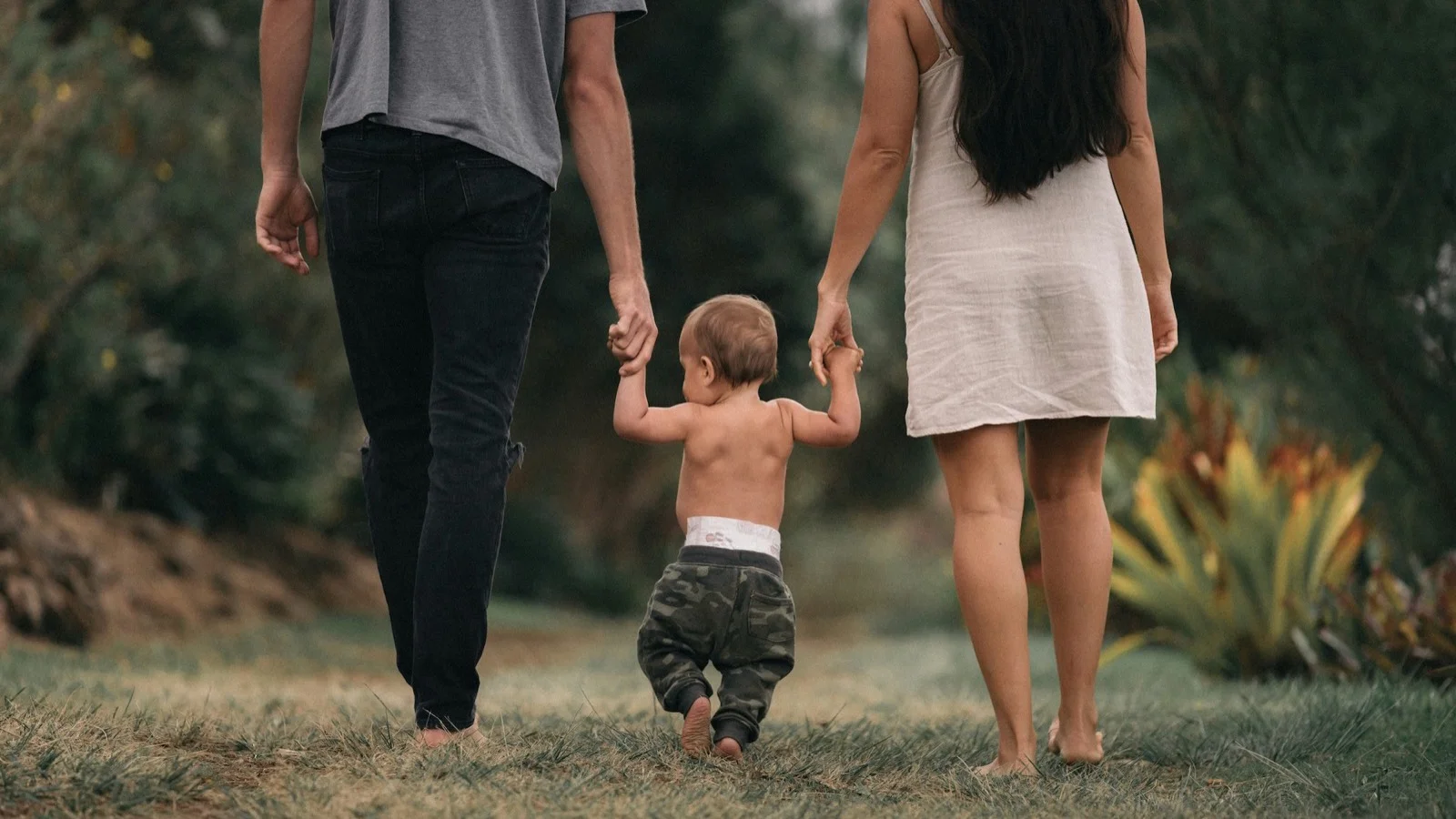 Two parents holding the hands of their baby learning to walk